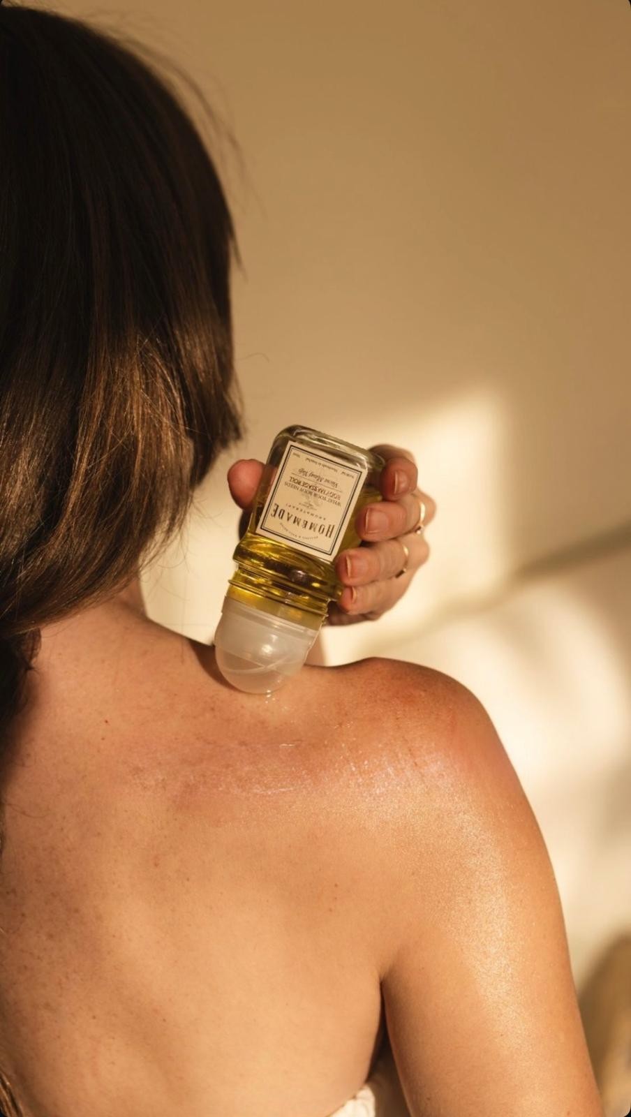 A person holding a golden oil bottle over their shoulder against a warm, sunlit background.